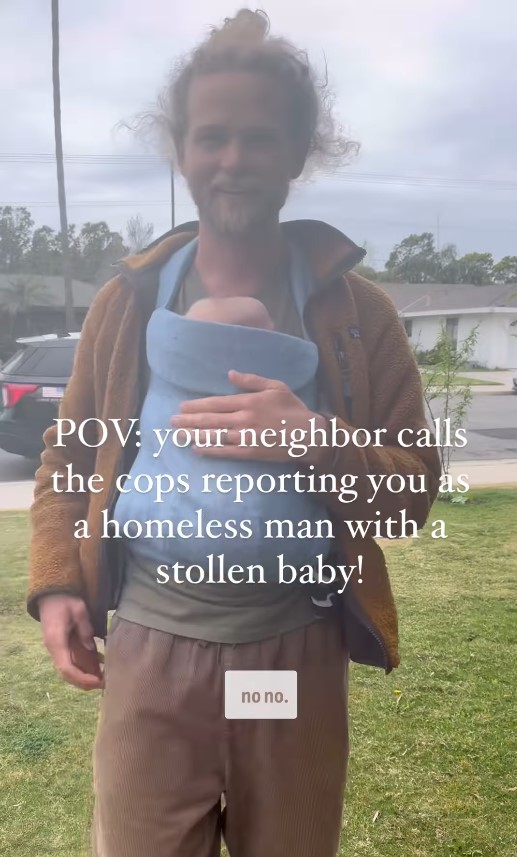 A man with a messy bun and beard stands on a suburban lawn, smiling while carrying a baby in a front-facing blue cloth baby carrier. He wears a brown fleece jacket, olive t-shirt, and loose brown pants. Overlay text reads: "POV: your neighbor calls the cops reporting you as a homeless man with a stollen baby!"
